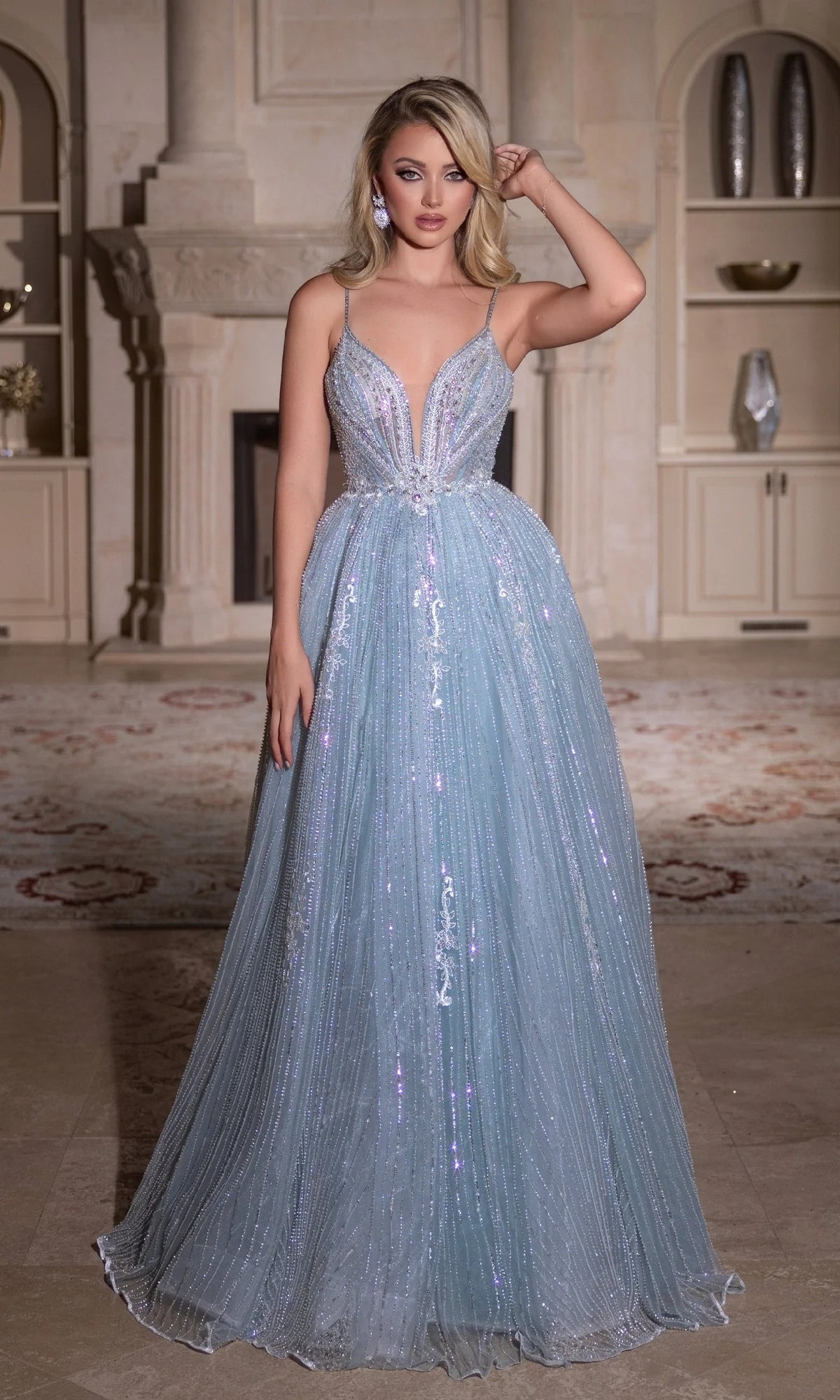 A woman stands indoors wearing the Long Prom Dress: Portia and Scarlett PS25310, a floor-length, light blue shimmery gown with a plunging neckline and thin straps. Shelves and a fireplace appear in the background.